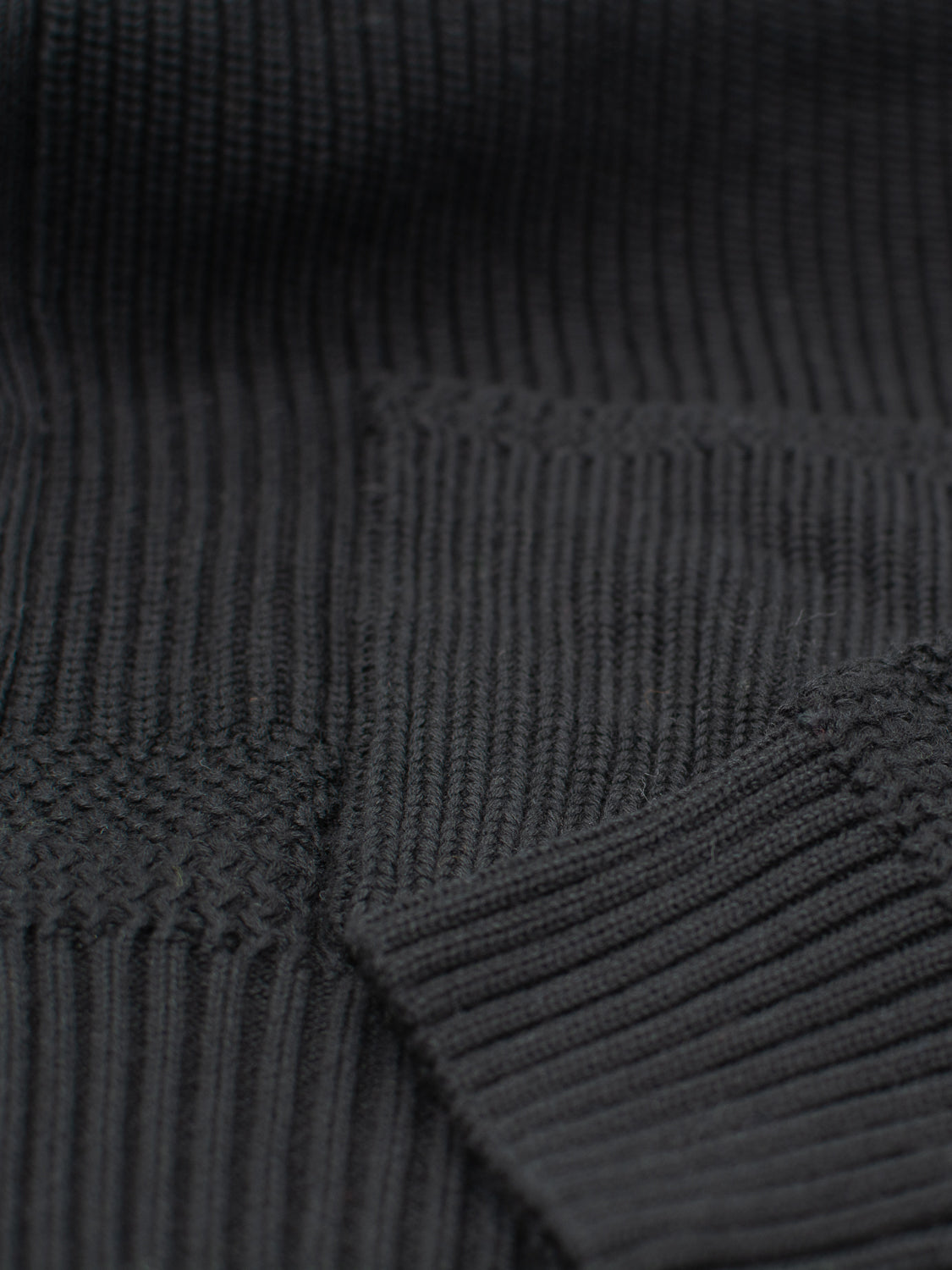 Close-up of a dark, ribbed knit fabric with varied textures, akin to the Helsinki Cotton Knitted Cardigan from ID Menswear. The mix of vertical and horizontal lines adds depth, and the thick material looks cozy, crafted from soft cotton.