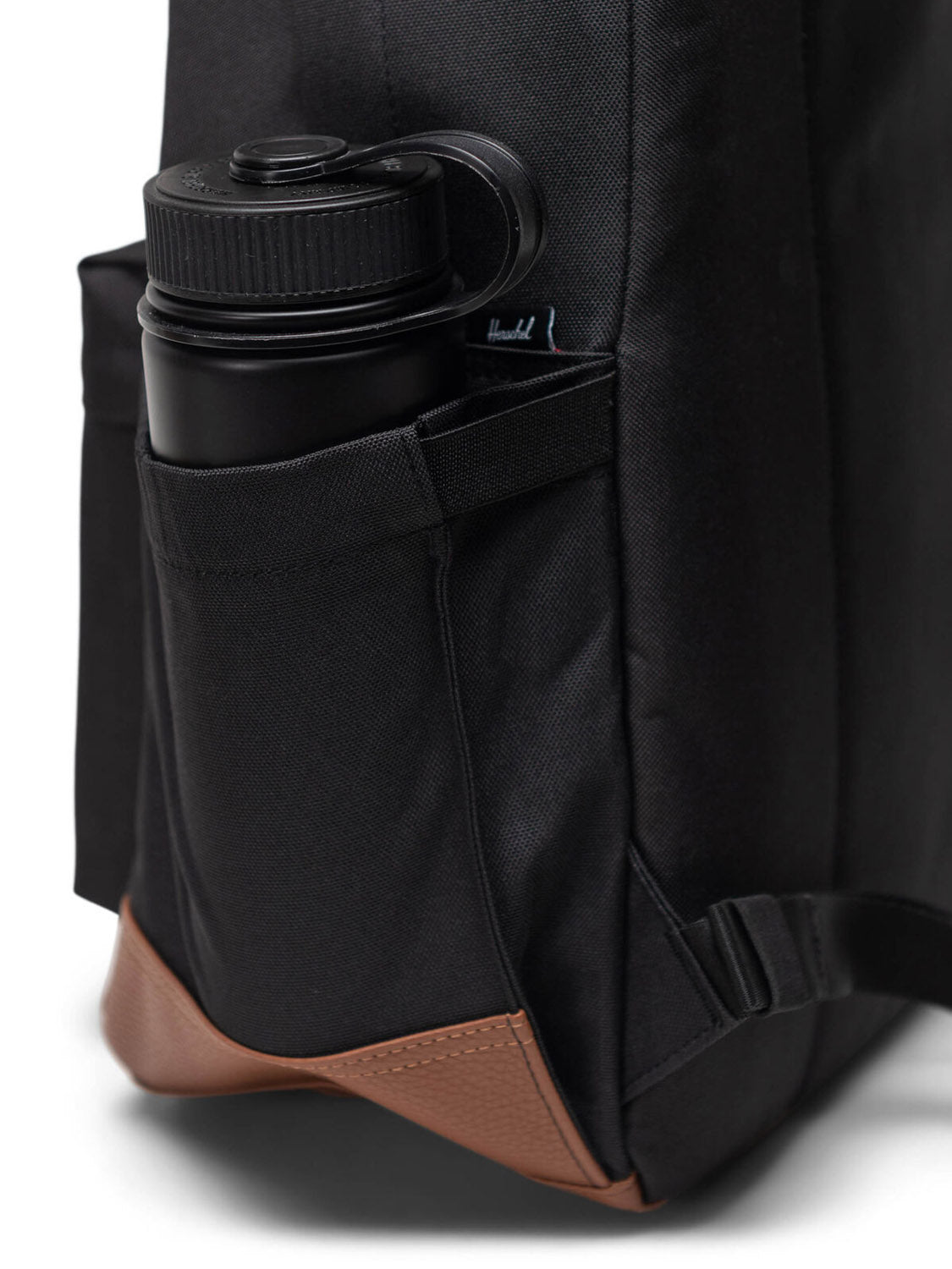 A black water bottle sits in the side pocket of a Herschel Heritage Backpack, which features brown leather trim on the bottom corner.