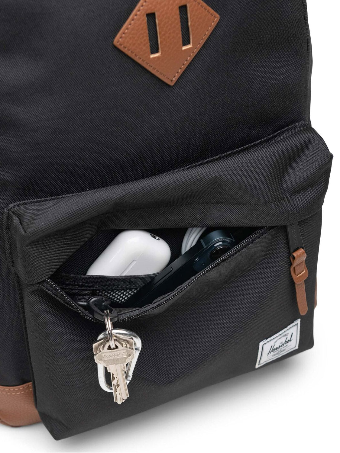 A close-up of the Herschel Heritage Backpack’s black front pocket, partially unzipped to show keys, a white AirPods case, and a dark item inside. Brown leather accents and the signature Herschel logo complete the sleek, functional design.