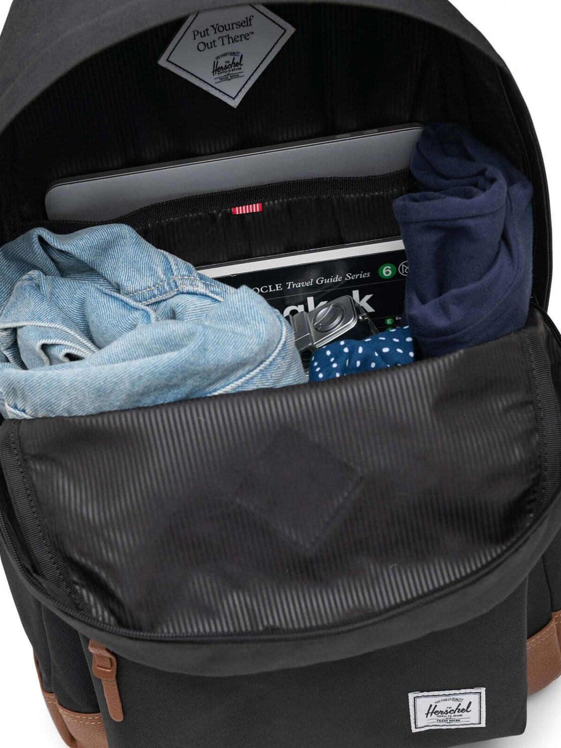 A Herschel Heritage Backpack in black is shown unzipped, revealing neatly packed clothing, a polka dot pouch, a travel guidebook, and a laptop inside the main compartment.