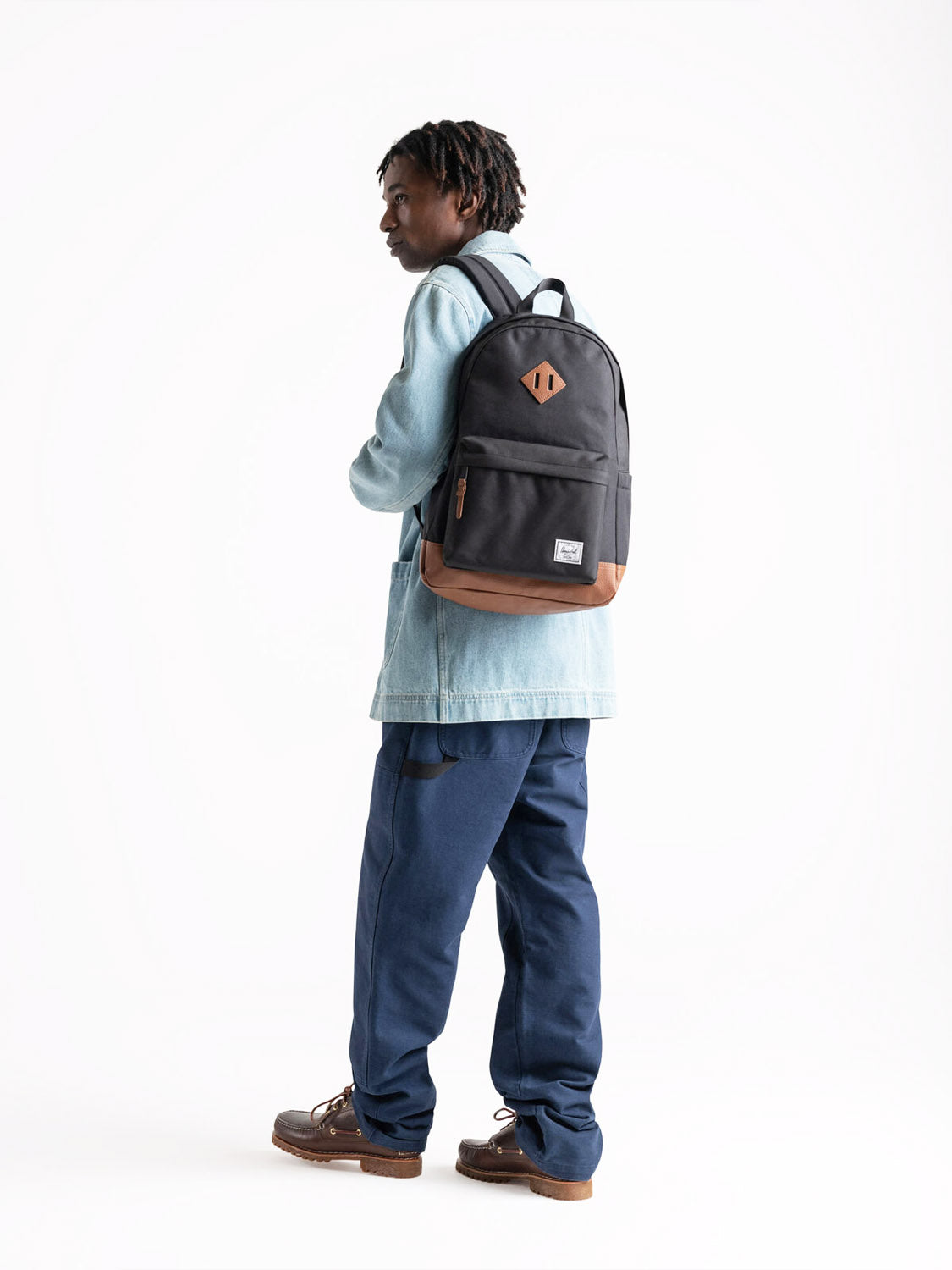 A person in a light blue denim jacket, blue pants, and brown shoes stands sideways on a plain white background, looking back while wearing the Herschel Heritage Backpack.