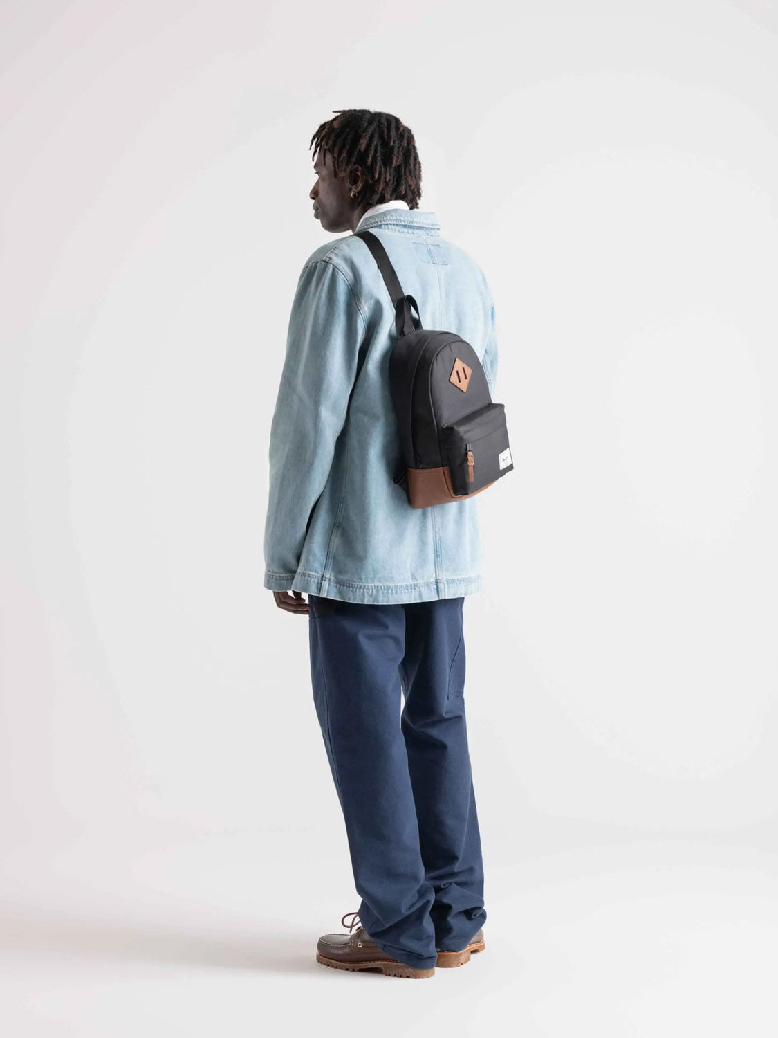 A person faces left against a plain white background, wearing a light blue denim jacket, navy pants, and brown shoes with the Herschel Heritage Shoulder Bag - 8L (black and brown, recycled polyester) on their back.