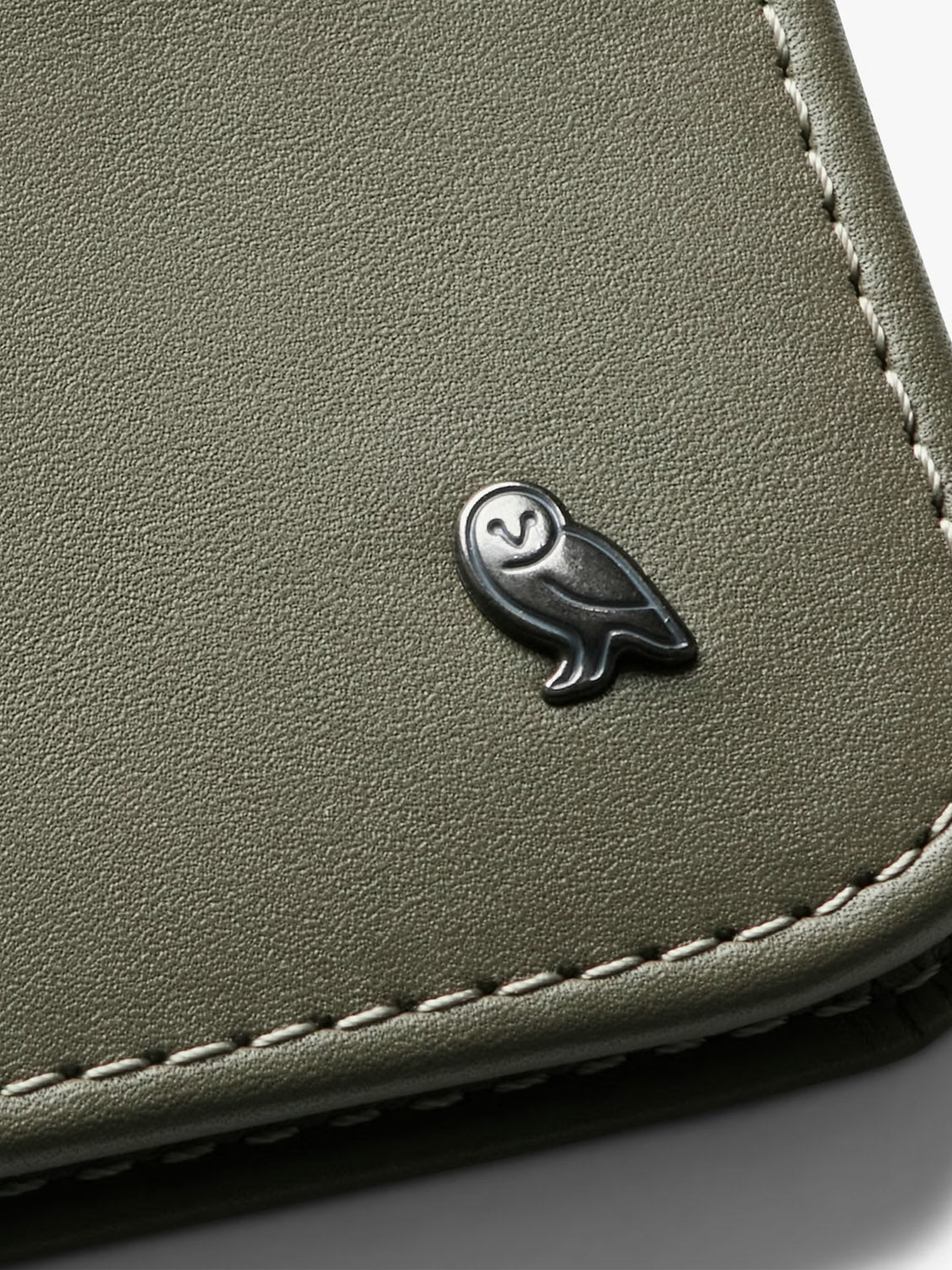 A detailed close-up of the Bellroy Hide and Seek Low Wallet in light olive green eco-tanned leather, featuring white stitching and a small metallic owl emblem on the corner.
