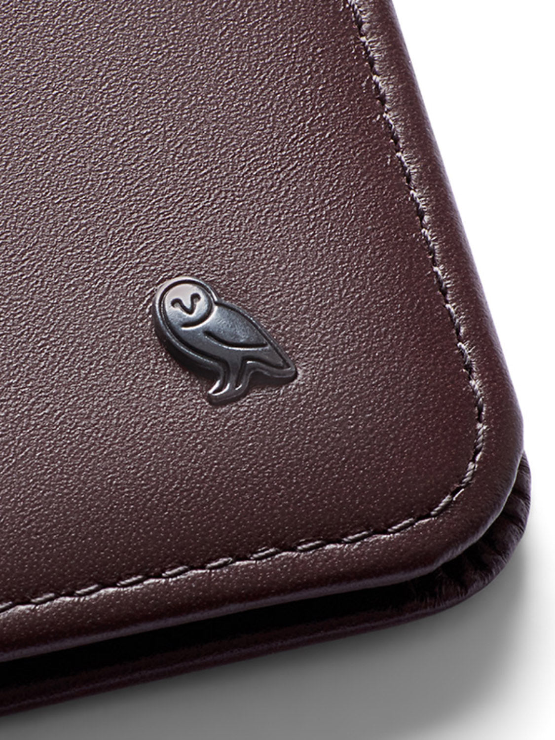 The Bellroy Hide and Seek Low Wallet features a close-up of its brown eco-tanned leather surface with a small embossed owl emblem in the corner, showcasing detailed edge stitching.