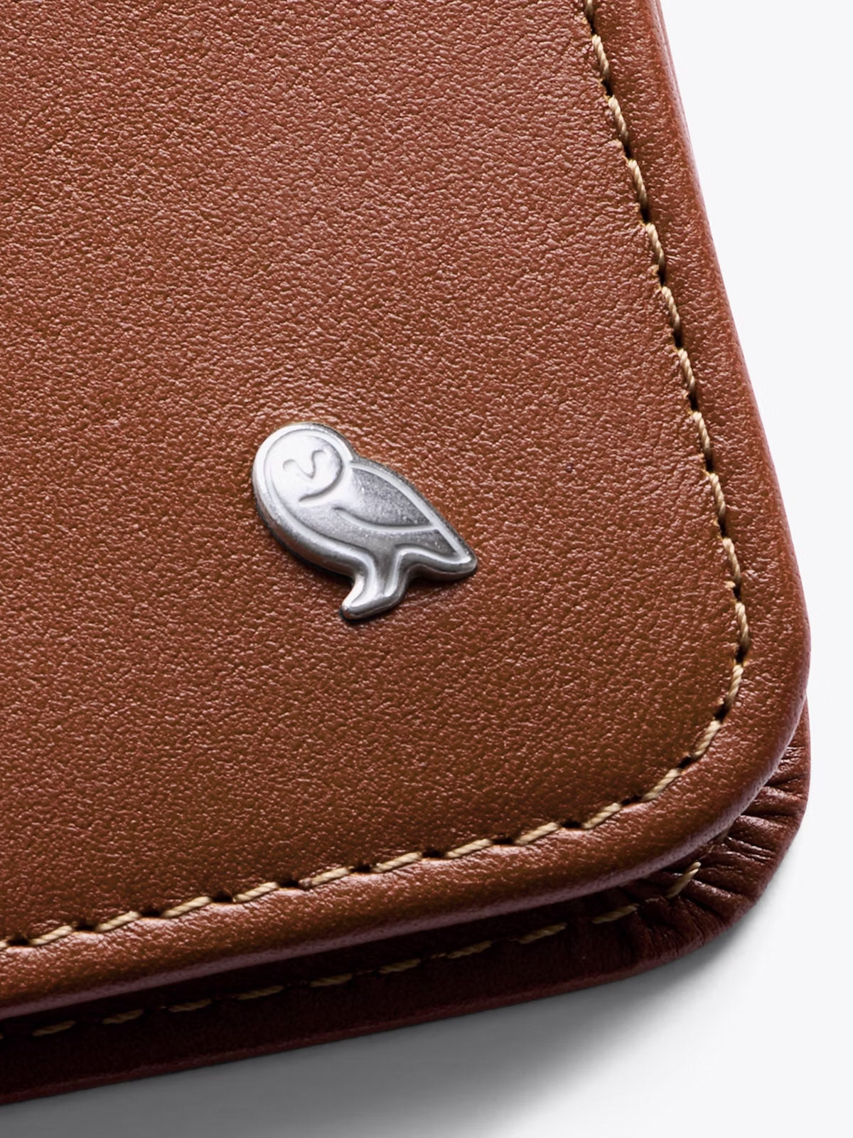 A close-up of the Bellroy Hide and Seek Low Wallet, made from brown eco-tanned leather, with precise stitching and a small silver owl emblem on the front.