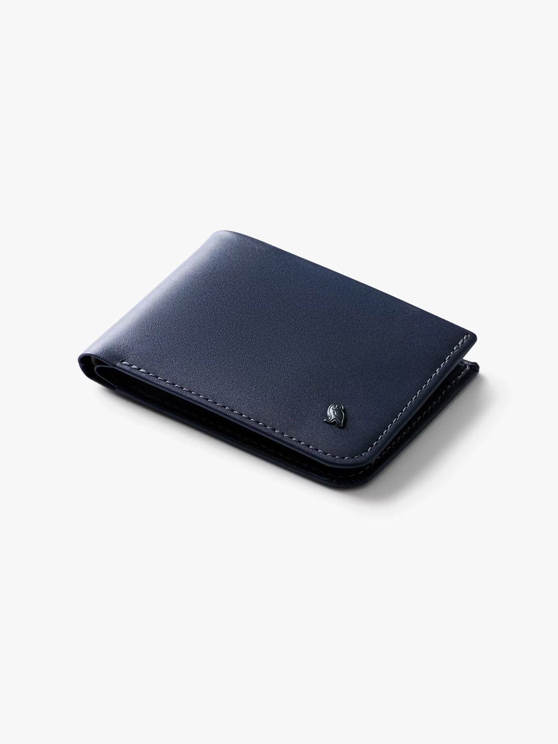 The Bellroy Hide and Seek Low Wallet is shown against a white background. It features a closed, navy blue slim design crafted from eco-tanned leather with subtle stitching and a small metallic emblem in the corner.