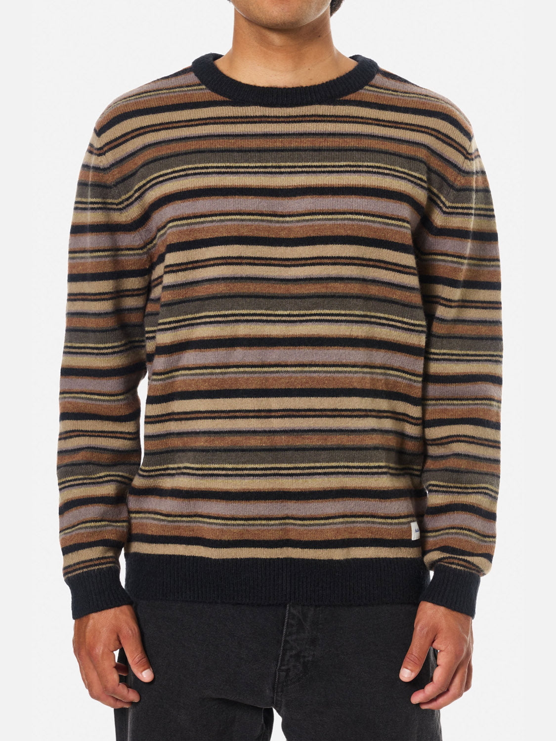 A person wears the Katin Hitch Sweater, made from ultra-soft blended yarn with a custom stripe pattern in brown, black, beige, and gray, paired with black pants on a plain white background.