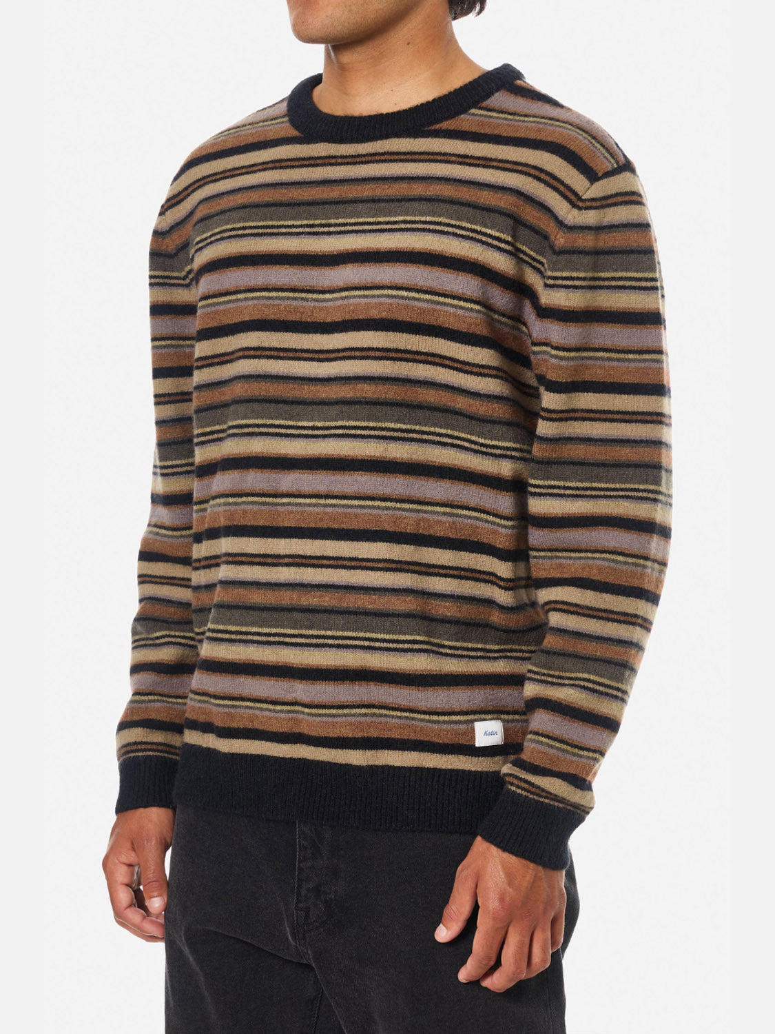 A person wearing the Katin Hitch Sweater, made from ultra-soft blended yarn with a custom stripe pattern in brown, tan, and black, paired with black pants. The person's face is not visible.