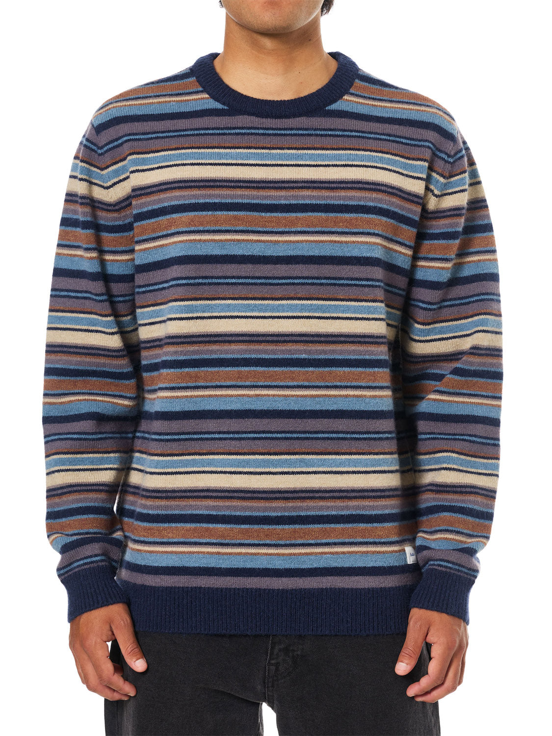 A person wears the Katin Hitch Sweater, made from ultra-soft blended yarn with a custom blue, brown, beige, and white stripe pattern, paired with black pants against a plain white background.