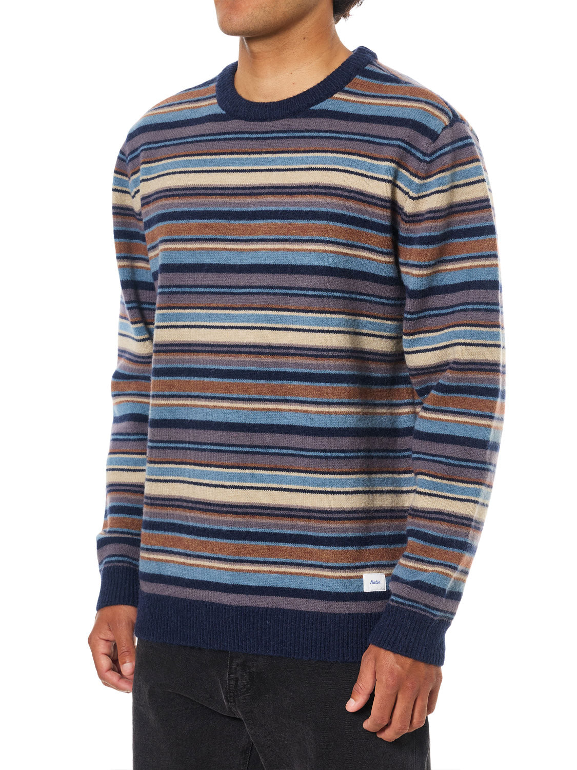 A model wears the Katin Hitch Sweater, featuring ultra-soft yarn and a custom stripe pattern in blue, brown, beige, and white, paired with black pants against a plain white backdrop.