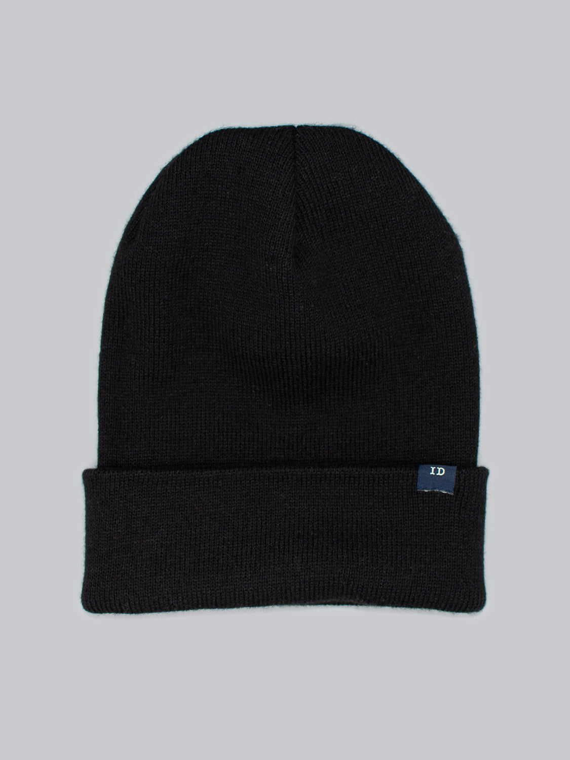 The ID Black Beanie by ID Menswear is crafted from 100% acrylic, featuring a folded brim and a small blue tag. Made in Brooklyn, its centered against a plain gray background.