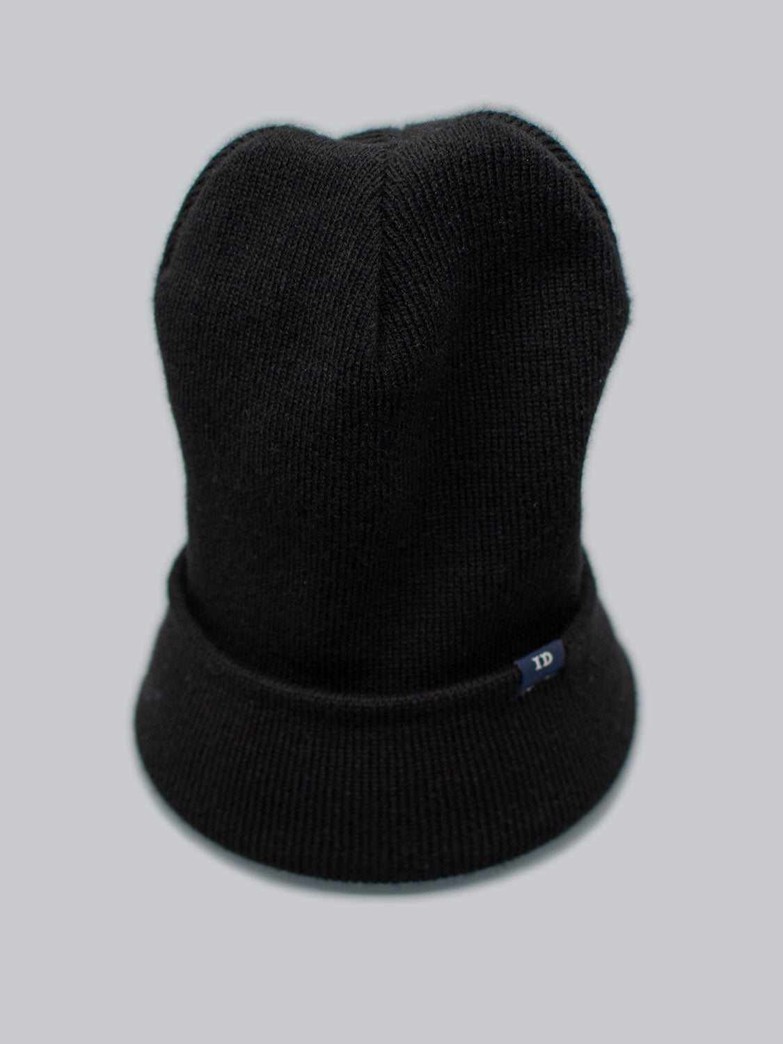 The ID Black Beanie from ID Menswear, made in Brooklyn with 100% acrylic, features a folded brim and a small dark tag displaying 1D. Its stylishly displayed against a plain gray background.