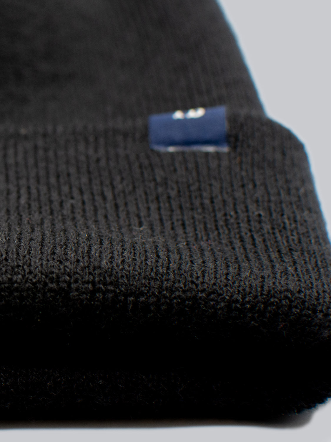 Close-up of the ID Black Beanie by ID Menswear, a folded black knit made in Brooklyn with a small blue tag. The soft, pale gray background highlights the texture of its 100% acrylic knit fabric.