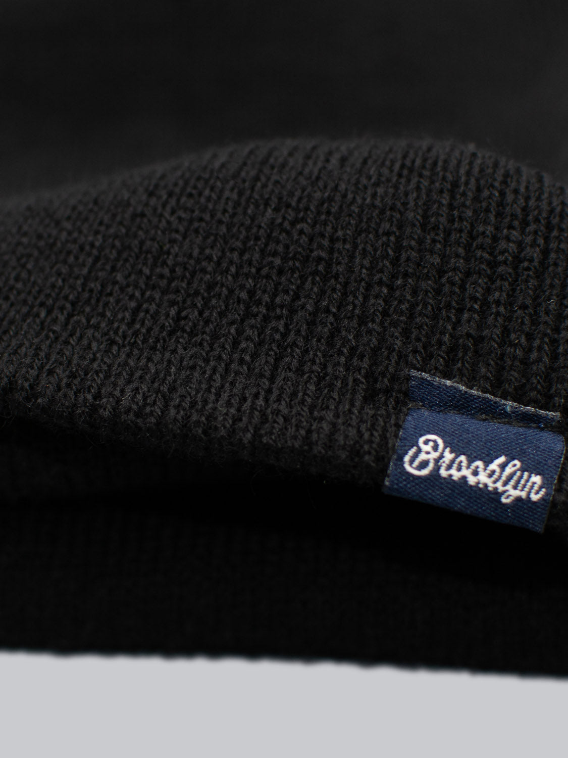 Close-up of an ID Black Beanie by ID Menswear, made in Brooklyn, showcasing a small navy tag on the brim with white cursive Brooklyn text against a gray background.