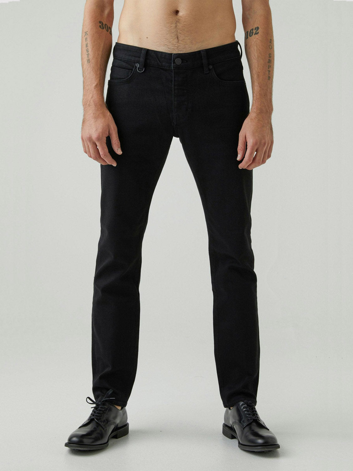 A shirtless person stands against a plain light background, arms relaxed at their sides, wearing Neuw's Iggy Skinny Perfecto black jeans with a slim waist and black dress shoes.
