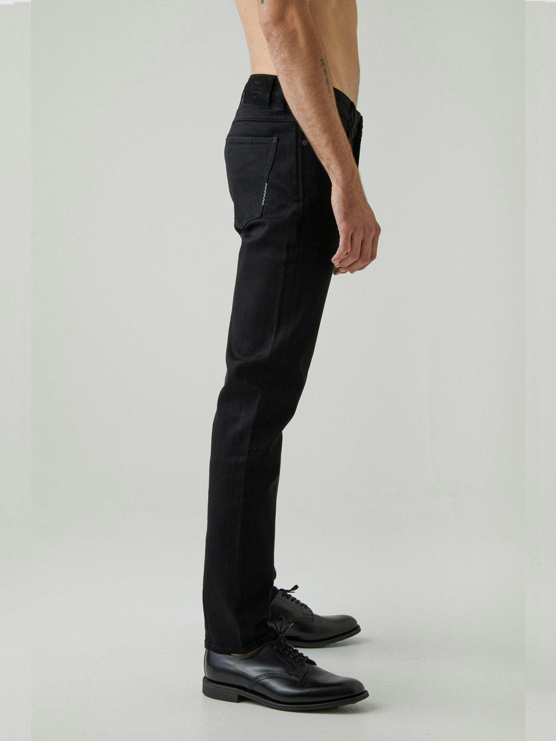 A shirtless person stands in profile wearing Neuw Iggy Skinny Perfecto black jeans and black dress shoes against a plain, light-colored background.