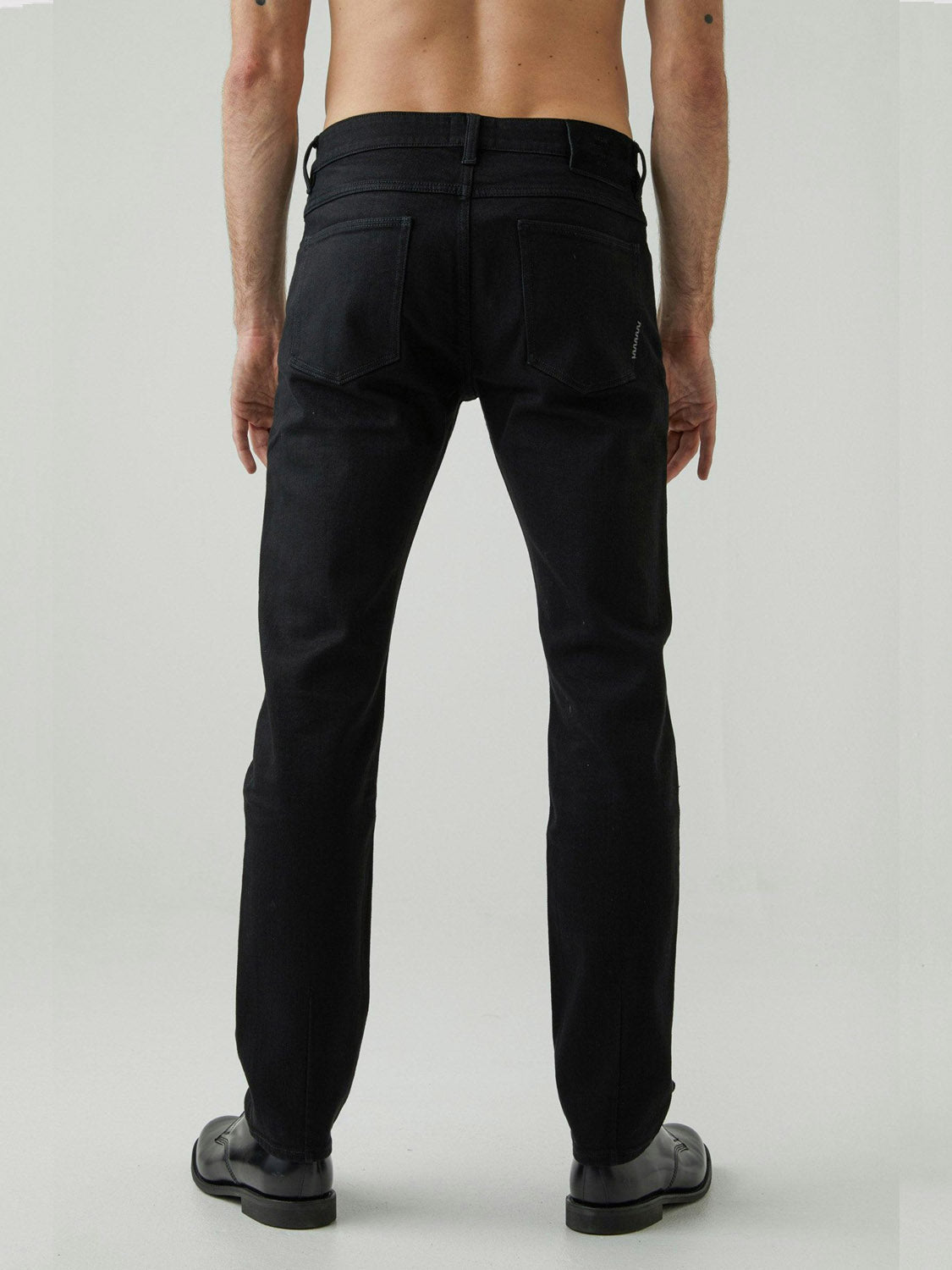 A shirtless person stands with their back to the camera, wearing Neuw's Iggy Skinny Perfecto jeans in black and black shoes against a plain light background.