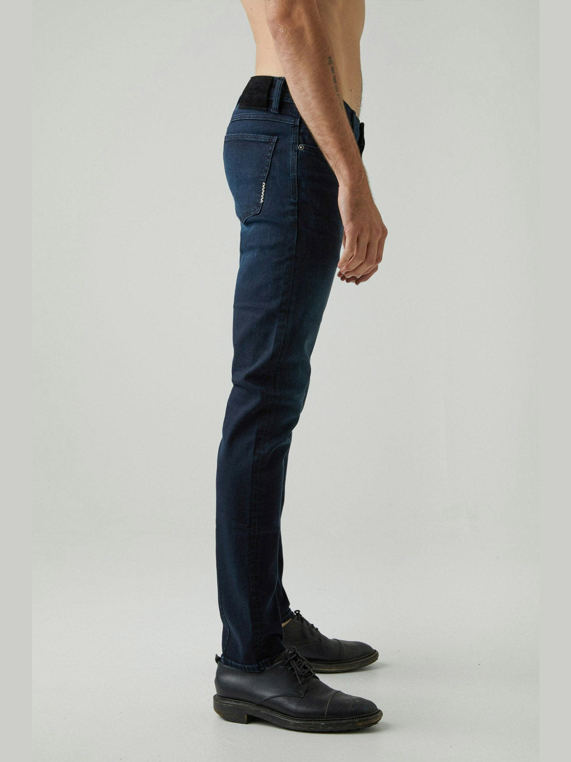 A shirtless man stands in profile against a plain light gray background, wearing Neuw's Iggy Skinny Polar jeans in dark blue with black dress shoes.