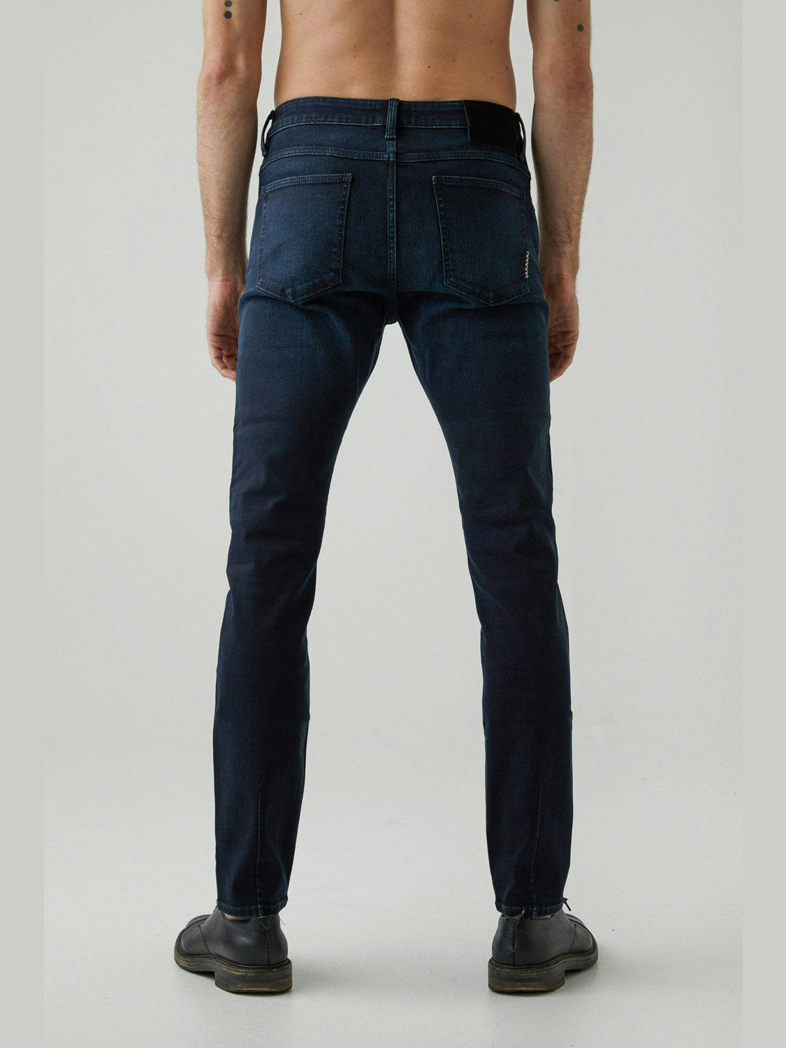 A shirtless person faces away, wearing Neuw Iggy Skinny Polar jeans with a skinny-tapered leg and dark shoes, set against a plain light background.