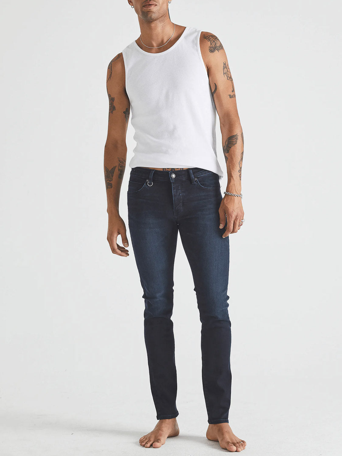 A tattooed man stands barefoot against a white background, wearing a fitted white tank top and Neuw Iggy Skinny Polar dark blue skinny-tapered jeans.