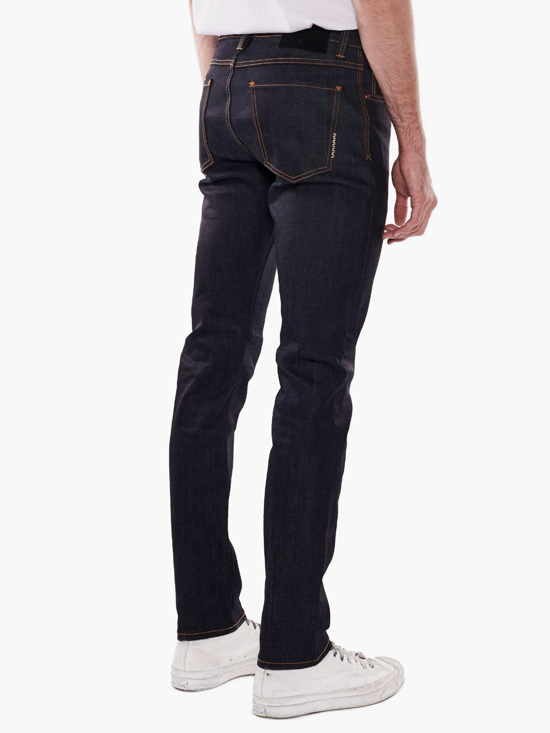 A man wearing Neuw’s Iggy Skinny Raw Stretch jeans, a white T-shirt, and white sneakers stands with his back to the camera and arms at his sides against a plain white background.