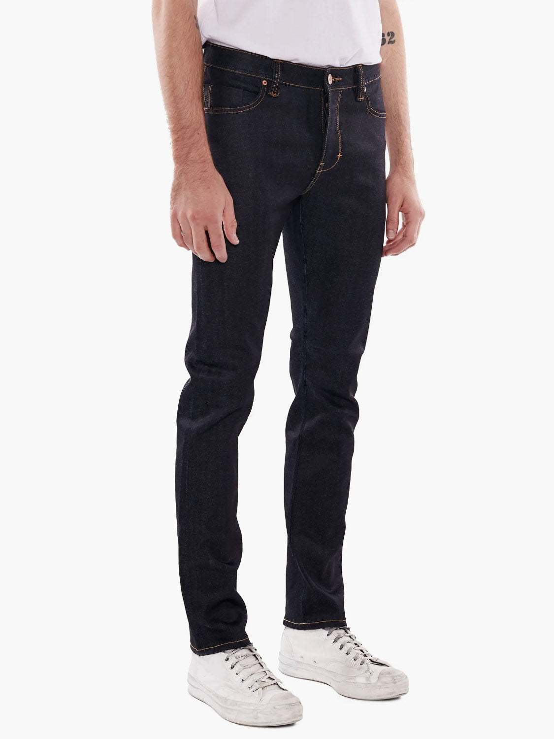 Only the lower half of a person is visible as they stand against a plain white background wearing Neuw Iggy Skinny Raw Stretch jeans, paired with a white t-shirt and white sneakers.