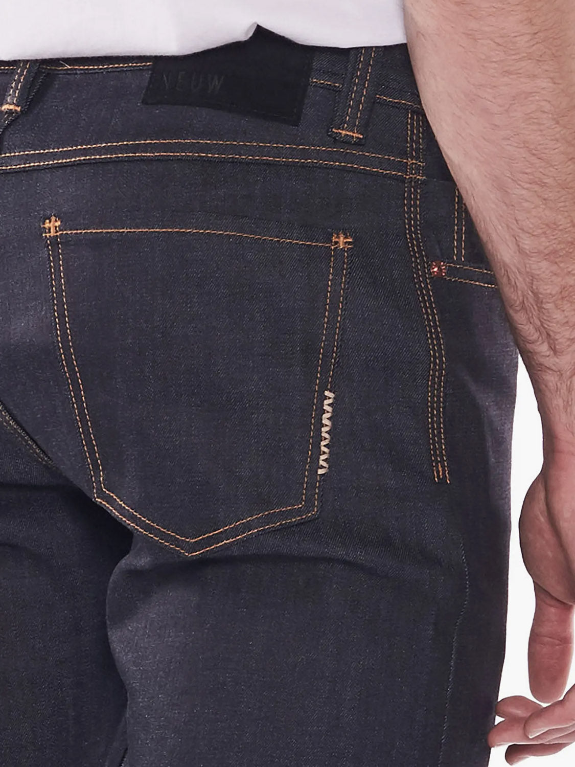 A close-up of the back of a person wearing Neuw’s Iggy Skinny Raw Stretch jeans with orange stitching, showing the rear pocket, part of their right arm and hand, and a partially visible waistband label.