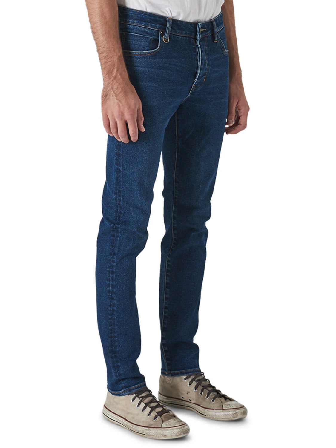 A person wears Neuw Iggy Skinny Zero Utility Blue jeans with a white shirt and worn white sneakers, standing against a plain white background. Only the lower half of the body is visible.