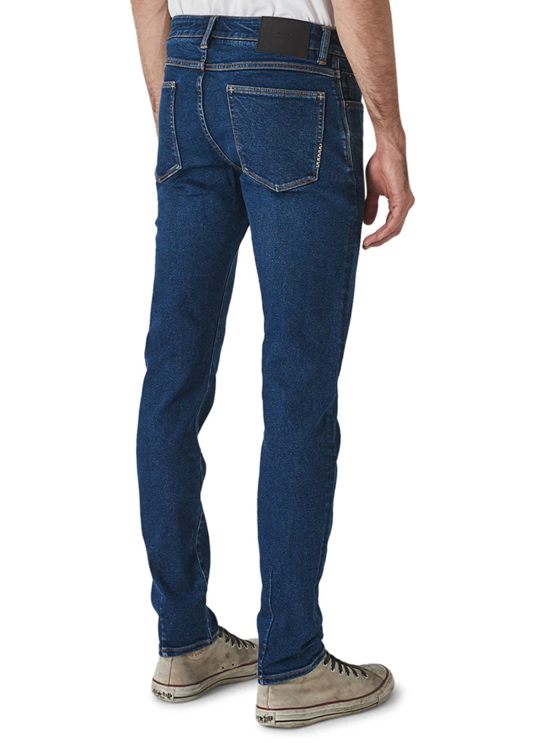 A person wearing Neuw Iggy Skinny Zero Utility Blue jeans and white sneakers is shown from the back. The jeans feature a slim waist, two back pockets with visible stitching, and a black patch on the waistband, against a plain white background.