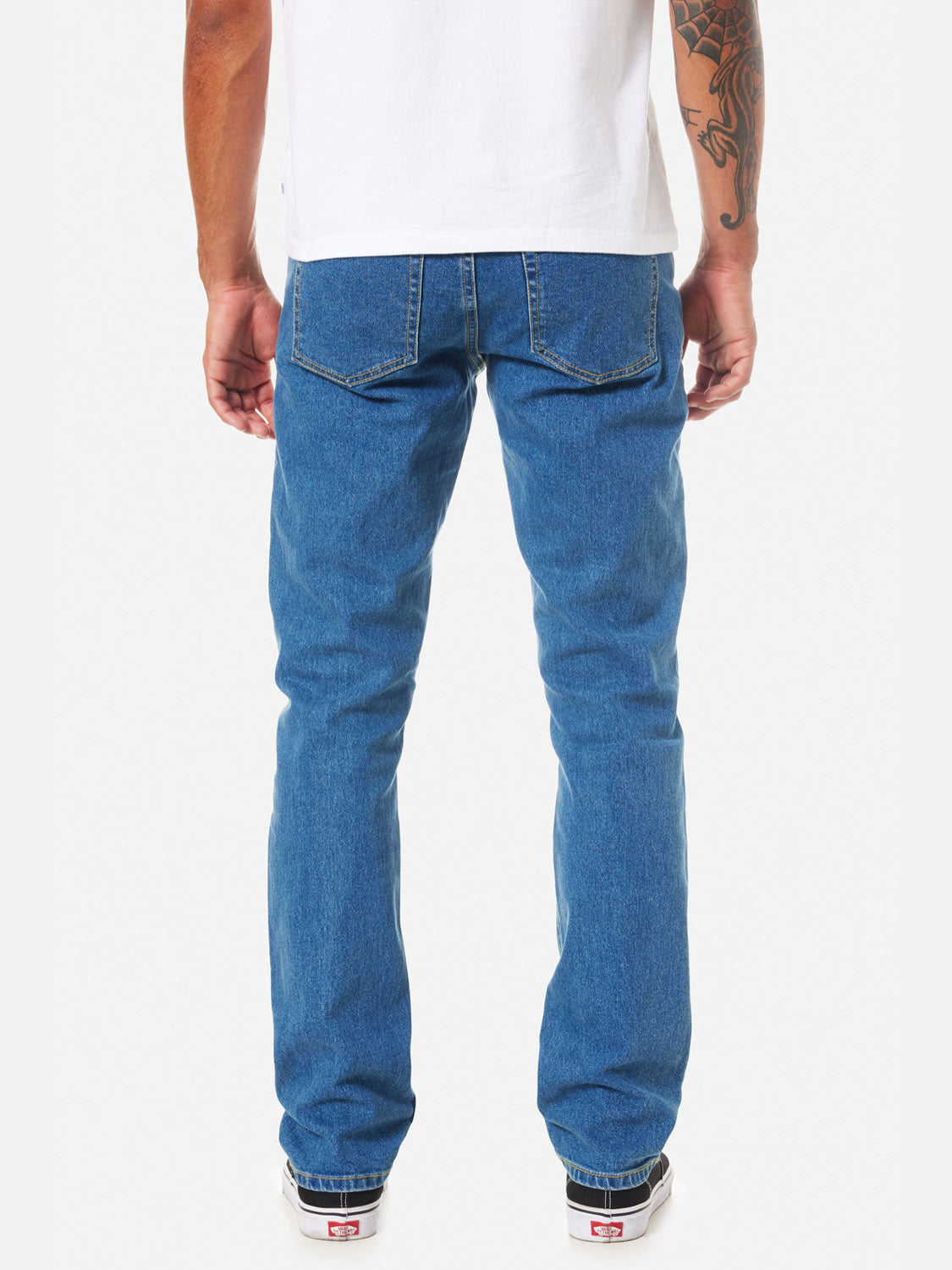 A person faces away on a plain white background, wearing a white T-shirt, Katin Jacob 71 Slim Straight Pants with classic 5-pocket styling, and white sneakers. One arm is tattooed.