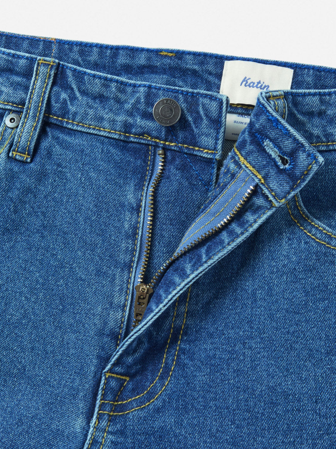 Close-up of the waist and fly area of Katin Jacob 71 Slim Straight Pants in blue denim, featuring yellow stitching, a partially open zipper, 5-pocket design, a metal button, and a waistband with an inner white Katin tag.