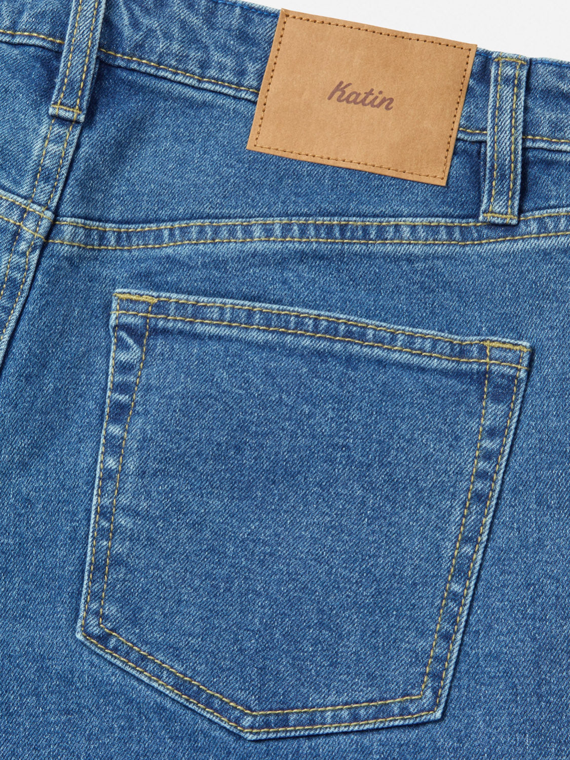 Close-up of the back pocket and waistband of the Katin Jacob 71 Slim Straight Pants in blue denim with yellow stitching. A brown leather patch above the pocket is embossed with the Katin brand. Features classic 5-pocket styling.