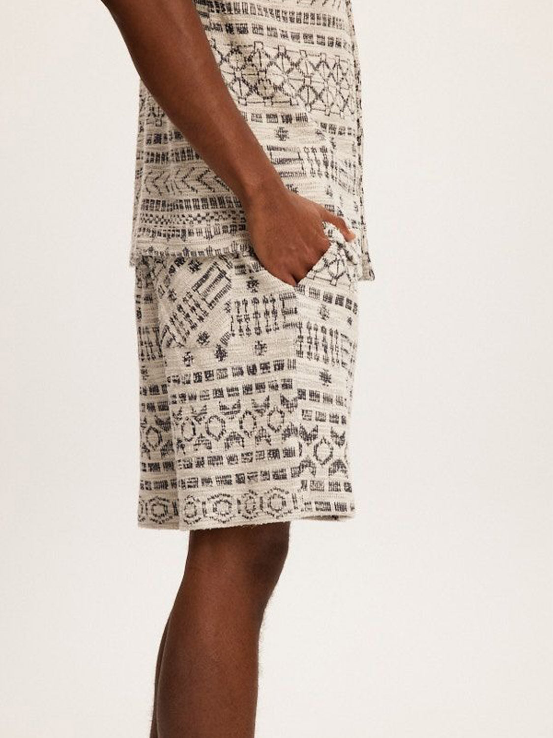 A person models the Kuwalla Jacquard Tribal Short set, featuring a detailed black and white tribal pattern. They stand sideways with a hand in their pocket, highlighting the high-quality fabric against a plain background.