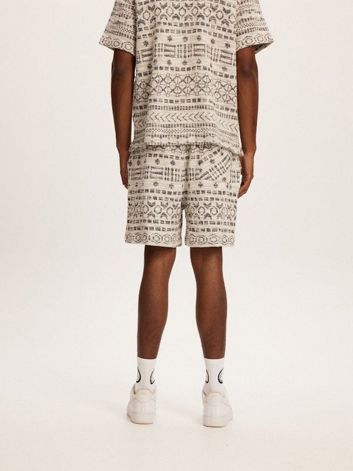 From behind, a person is spotted against a plain backdrop, wearing Kuwallas Jacquard Tribal Short set with white sneakers and socks. The ensemble is crafted from high-quality fabric, showcasing geometric designs in neutral tones for both the shirt and shorts.