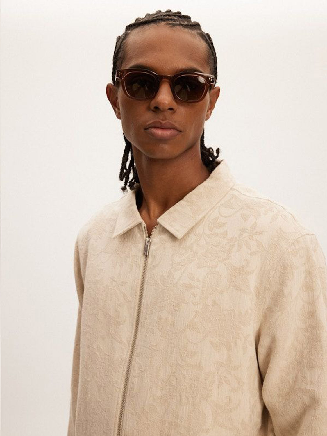 A person with braided hair and dark sunglasses wears a Kuwalla Jacquard Linen Shacket—relaxed fit with a subtle floral pattern—against an off-white background, perfect for transitional weather.
