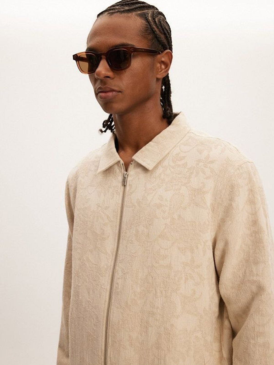 A person with braided hair wears rectangular sunglasses and a beige Kuwalla Jacquard Linen Shacket, featuring a subtle floral pattern and relaxed fit with a collar and front zipper, ideal for transitional weather as they stand against a plain light background.