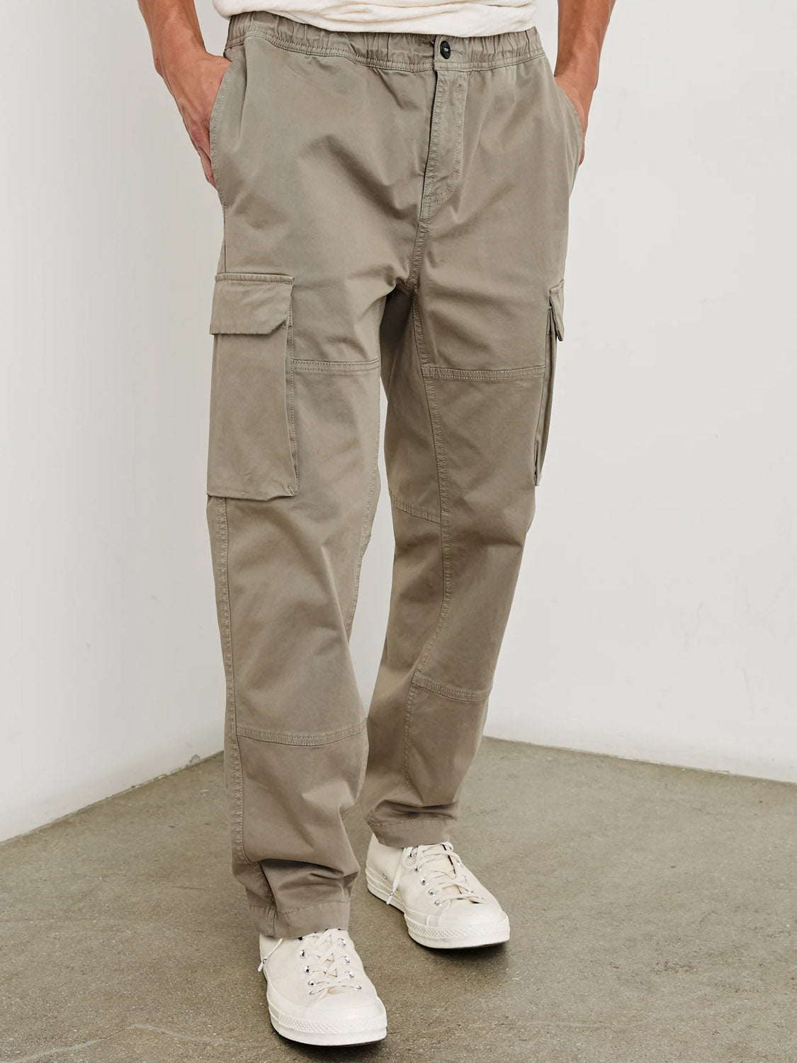 A person wears Rails Jaiden Cargo Pants in light khaki, featuring side pockets, an elastic waistband, and a relaxed fit, paired with white sneakers and a white shirt on a concrete floor against a plain white wall.