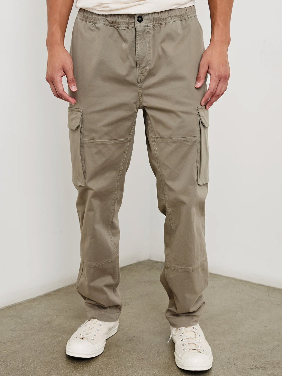 A person wearing Rails Jaiden Cargo Pants in light brown with side pockets and white sneakers stands against a light wall on a concrete floor, showing only the lower half of their body.