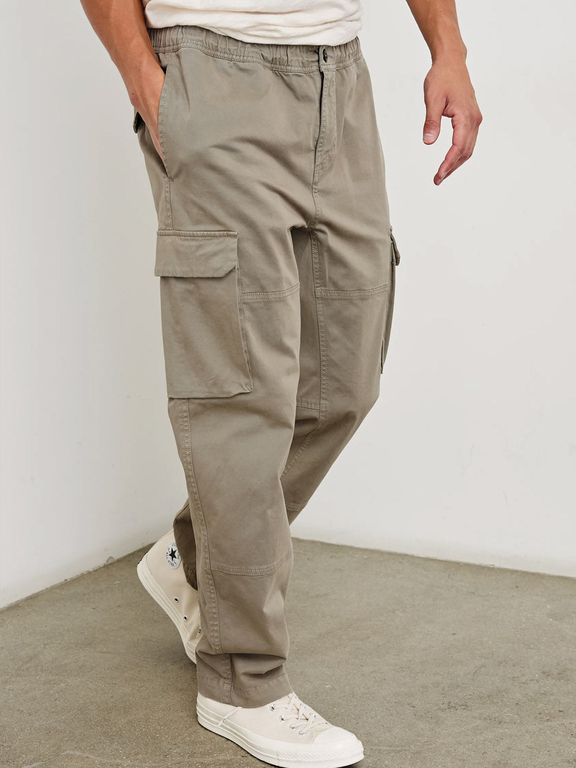 Someone stands against a plain wall and concrete floor, wearing Rails Jaiden Cargo Pants in light beige with side pockets and white sneakers, one hand tucked into a pocket.