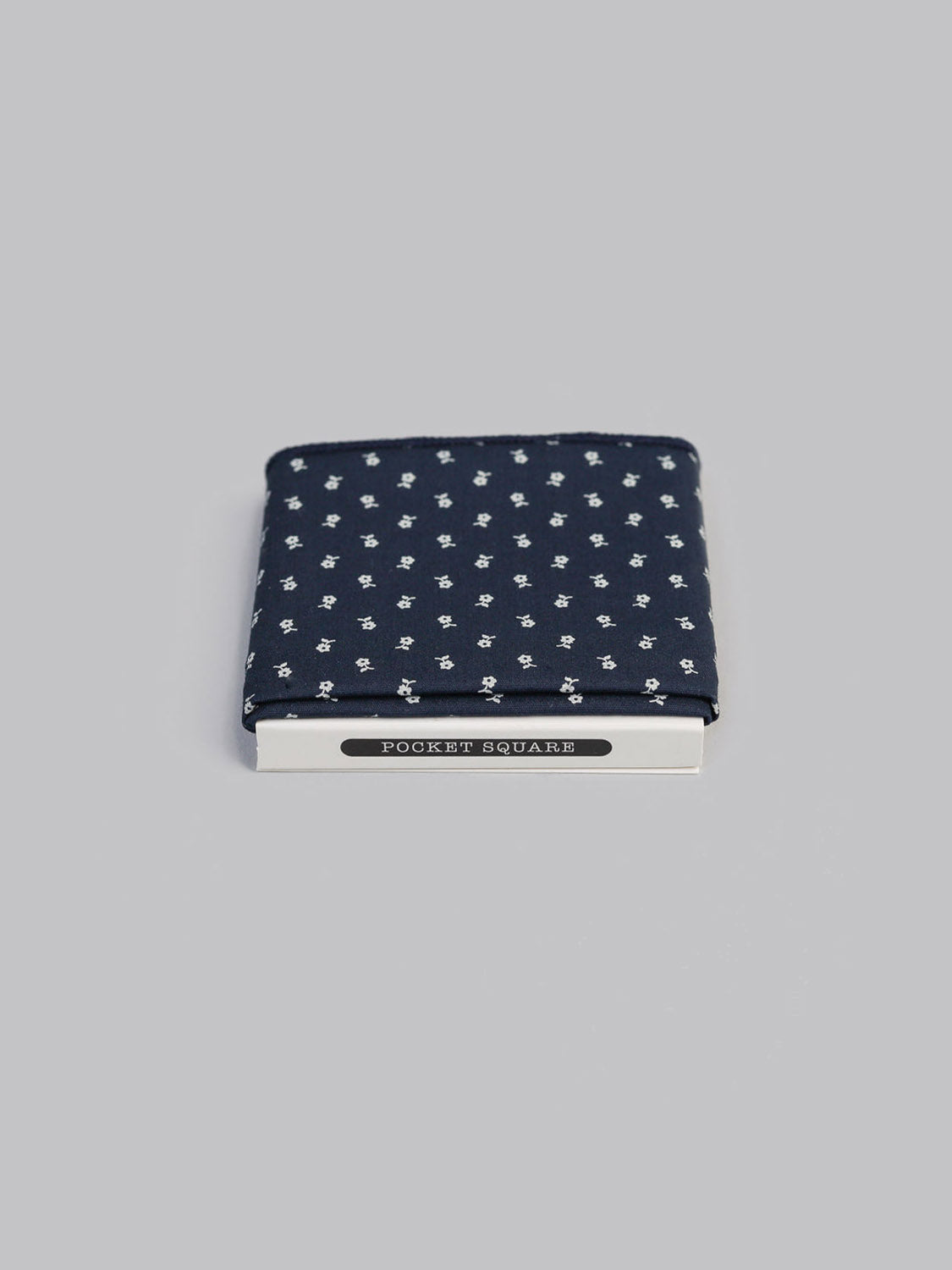 The Jaipur Pocket Square by ID Menswear, handmade in Brooklyn from 100% cotton navy blue fabric with a white floral pattern, comes neatly folded in white POCKET SQUARE packaging against a plain gray background.