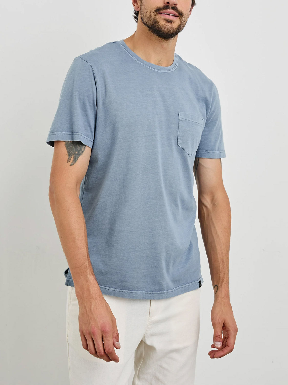 A person is wearing the Rails Johnny Tee, a light blue classic fit T-shirt with a chest pocket, paired with off-white pants. Made from super soft cotton modal fabric for added comfort, they have a tattoo on their right forearm and are standing against a plain white background.