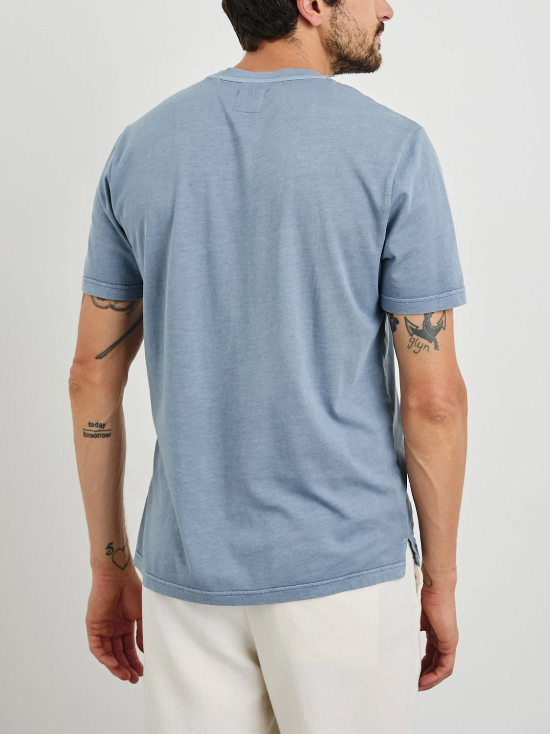 A man with short dark hair and a beard wears the Rails Johnny Tee in light blue, featuring a vintage wash. He pairs it with white pants, complementing his arm tattoos. Hes standing against a plain white background, showcasing an effortlessly cool style.