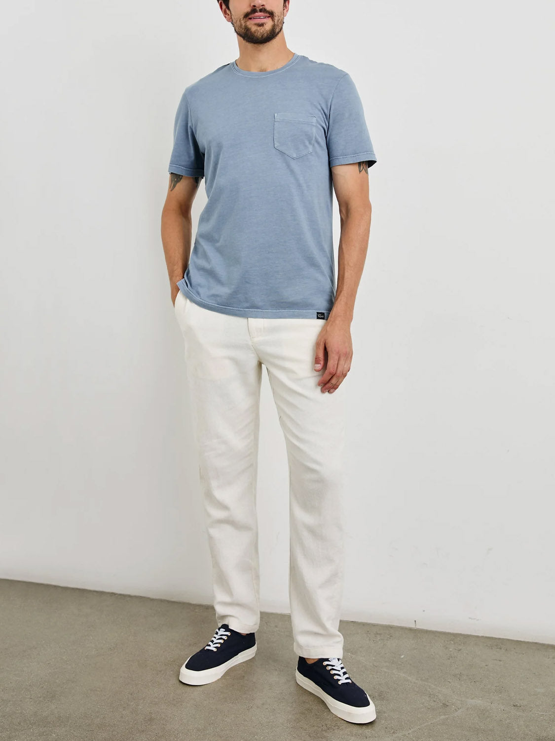 Against a plain white wall, a person wears a Rails Johnny Tee in vintage light blue wash, white pants, and black sneakers with white soles. One hand is in their pocket while the other hangs by their side. The floor is gray.