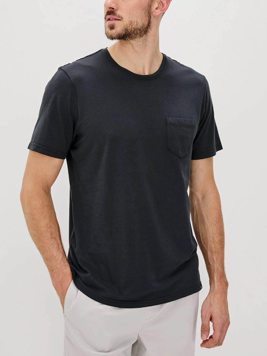 A man wears the Rails Johnny T-Shirt, a plain black soft cotton jersey short-sleeve tee with a chest pocket, paired with light-colored pants, standing against a white background.