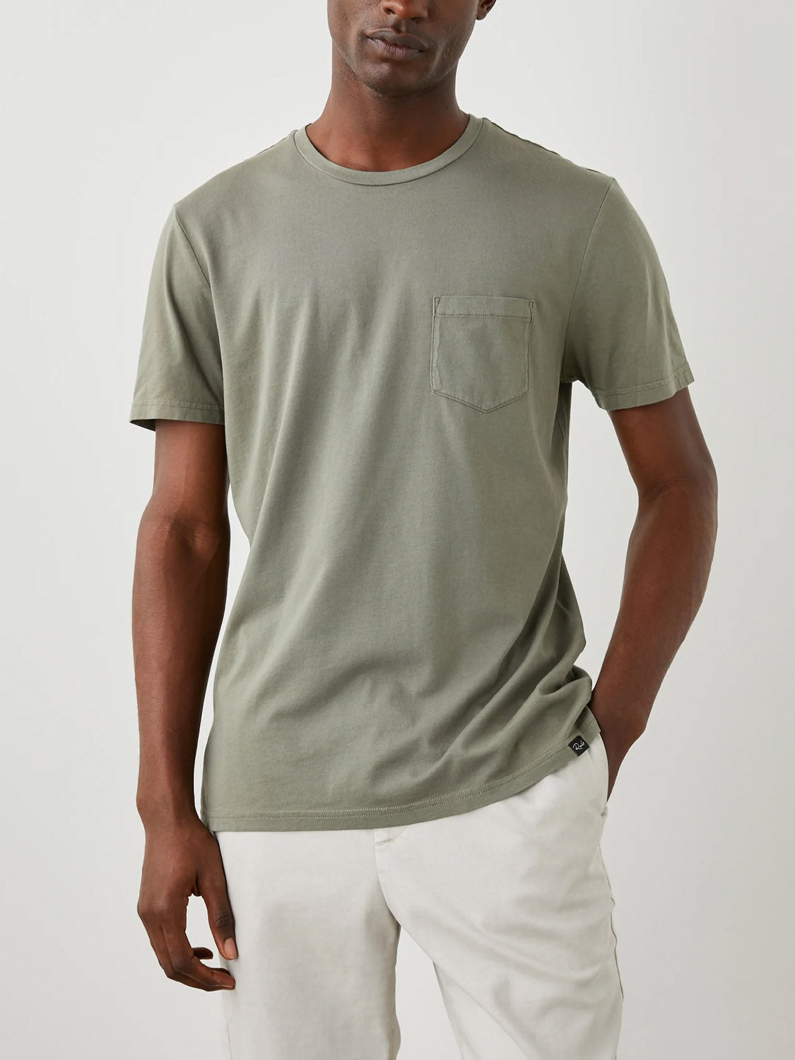 A person models the Rails Johnny Tee, an olive green short-sleeve T-shirt made from supremely soft cotton modal with a vintage wash effect. Featuring a chest pocket and relaxed fit, the tee pairs easily with light-colored pants, as one hand rests in their pocket against a plain background.