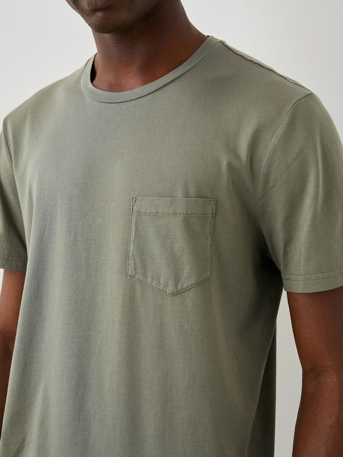 A person wears the olive Rails Johnny Tee, featuring super soft cotton modal, a left-side pocket, and a classic fit with a vintage wash. Only their lower face and neck are visible against a plain light-colored background.