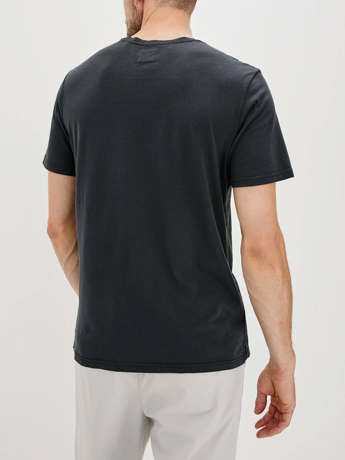 A man, seen from behind against a plain light background, is wearing the Rails Johnny T-Shirt, a plain black soft cotton jersey short-sleeve tee, paired with light gray pants.