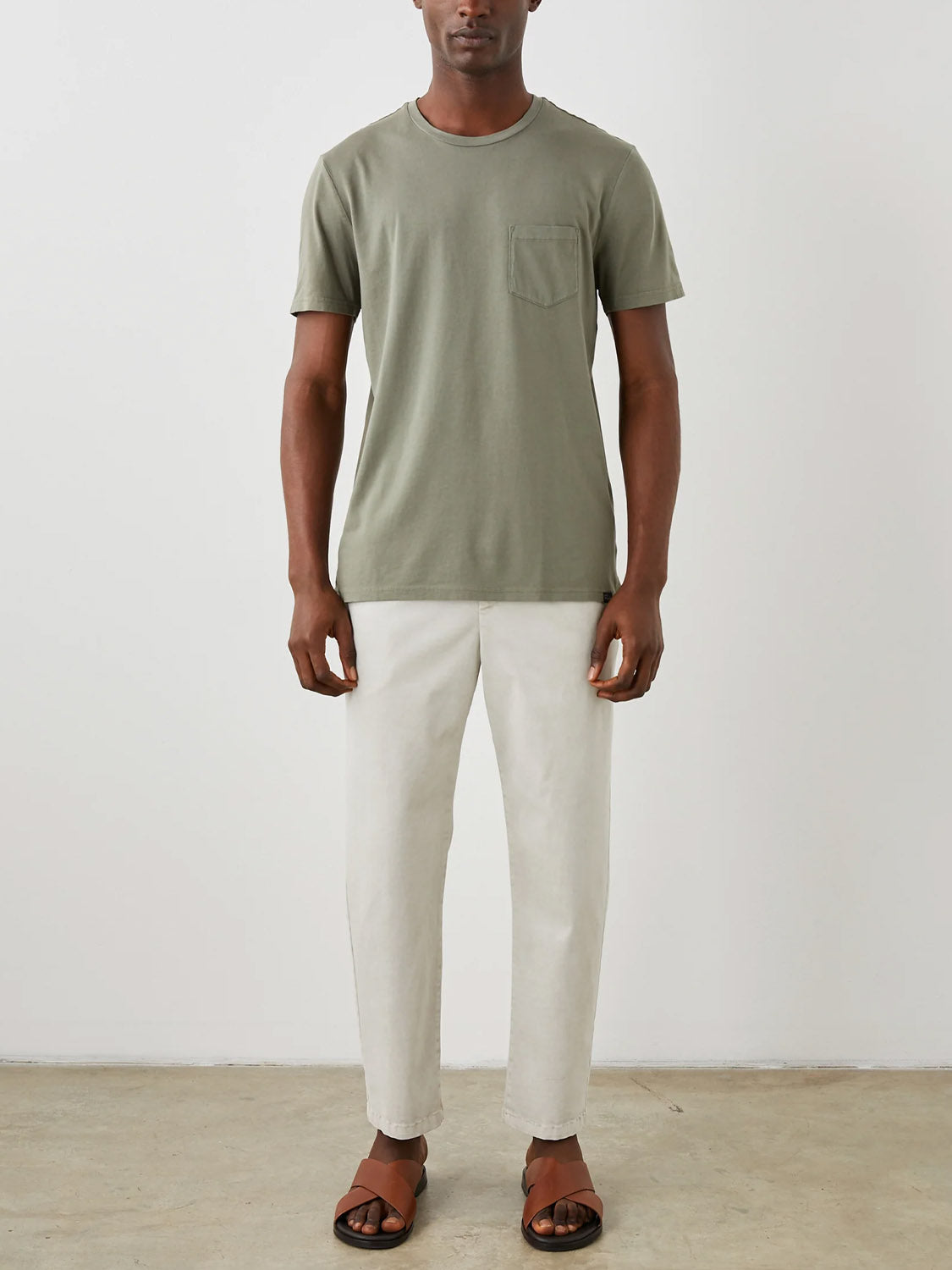 A person stands against a plain background, wearing the Rails Johnny Tee in olive green with a classic fit and vintage wash, paired with light beige pants and brown sandals. Made from super soft cotton modal for added comfort, the setting is neutral, possibly indoors on a light-colored floor.