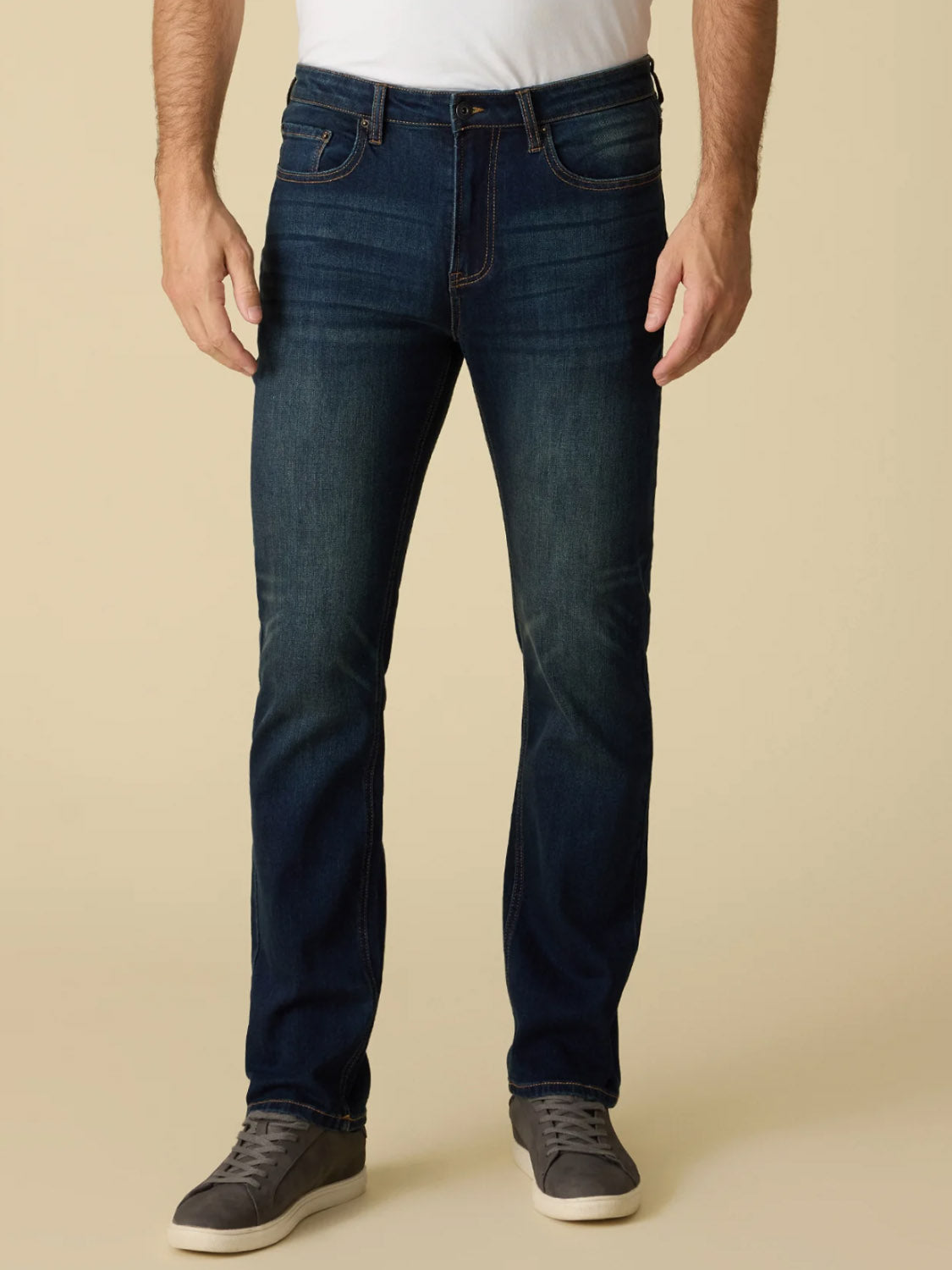 A man wearing Flag and Anthem Jonesville Cooling Jean-Nashville Straight stands against a beige background. He pairs the jeans with a white t-shirt and gray sneakers. His hands are by his sides, and his head is not visible in the image.