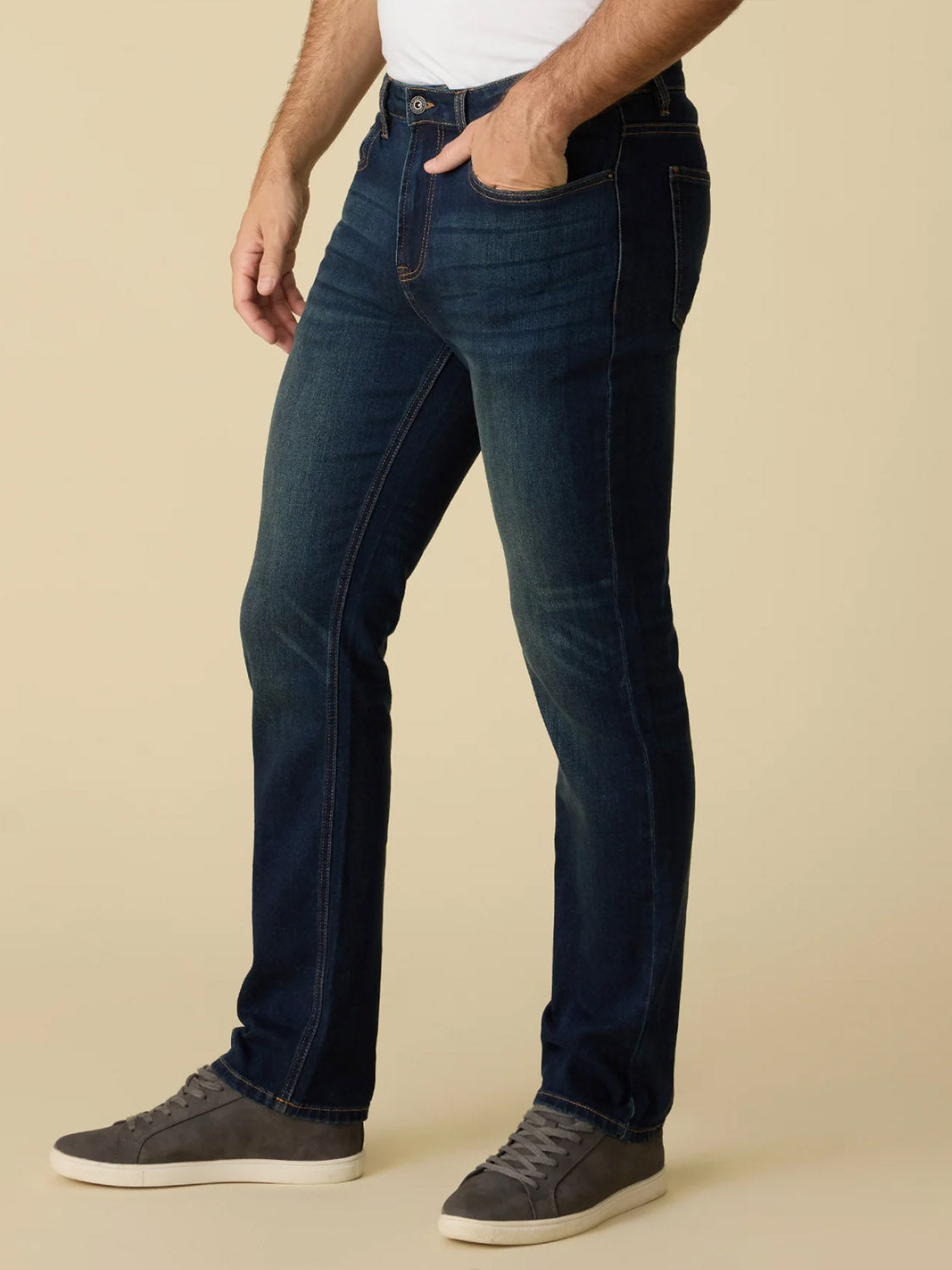 Wearing Flag and Anthem's Jonesville Cooling Jean-Nashville Straight in dark blue, paired with a white shirt and gray sneakers, a person poses with one hand in their pocket against a beige background, showing only their lower half.